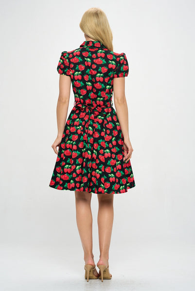 Model wearing a cotton short sleeve flared dress with a notched v-neck collar, princess seaming, puffed cap sleeves, a self removable sash belt, and a flared above the knee skirt. Dress is black with a red and green strawberry pattern. Shown from the back