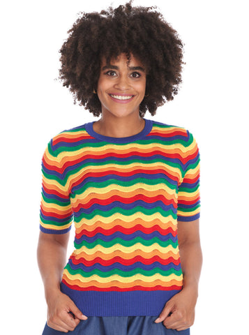 A model wearing a short sleeved sweater with a round neckline and elbow length sleeves in an all over knit in wavy rainbow pattern with small pointelle details at the top of each wave. The sweater has bright blue ribbed collar, cuffs, and bottom band. Shown from the front untucked 