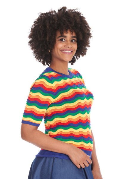 A model wearing a short sleeved sweater with a round neckline and elbow length sleeves in an all over knit in wavy rainbow pattern with small pointelle details at the top of each wave. The sweater has bright blue ribbed collar, cuffs, and bottom band. Shown from a three quarter angle untucked