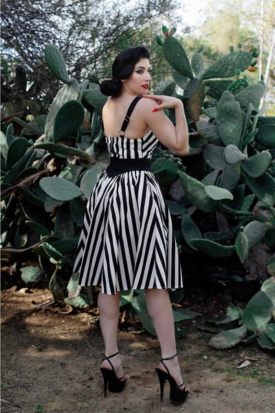 A model wearing a playsuit and skirt set made of stretch black and white cotton. The playsuit has a sweetheart neckline, princess seaming, and wide black adjustable straps. The skirt is a just below the knee swing skirt with a solid black yoke style waistband. Shown from the back