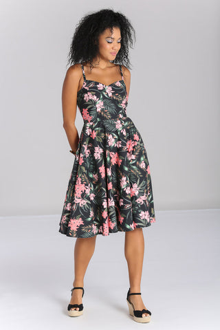Model wearing a sleeveless 1950’s style sundress in a stretch cotton fabric. All-over print on a black background of pink tropical flowers and green foliage. Dress has a sweetheart neckline, princess seaming, adjustable thin straps, and a full below the knee skirt with pockets. Seen from the front 