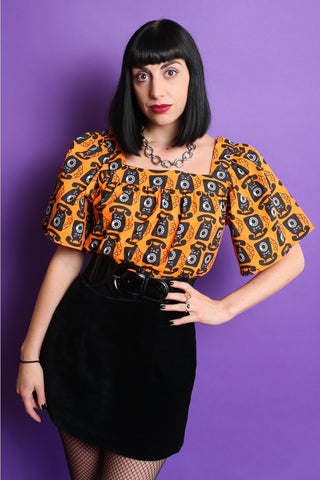 A model wearing a lightweight cotton peasant style top with an all over pattern of a black cat with rotary phone features on a bright orange background. It has a square neckline and short bell sleeves ending just above the elbow. Seen tucked in from the front