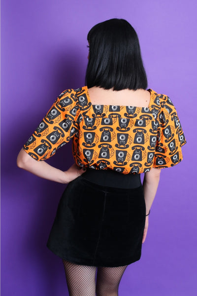 A model wearing a lightweight cotton peasant style top with an all over pattern of a black cat with rotary phone features on a bright orange background. It has a square neckline and short bell sleeves ending just above the elbow. Seen tucked in from the back