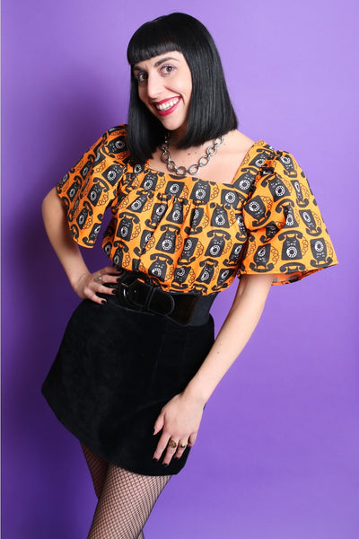A model wearing a lightweight cotton peasant style top with an all over pattern of a black cat with rotary phone features on a bright orange background. It has a square neckline and short bell sleeves ending just above the elbow. Seen tucked in from a three quarter angle