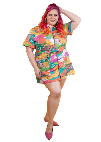 A model wearing a stretch cotton mini dress with a rounded wide collar, short sleeves, and a bright purple plastic zipper running down the length of the dress. It has an intricate multicolored psychedelic pattern including pinwheels, apples, hands, birds, and other illustrations in pink, yellow, orange, blue, green, and white. Dress comes with a matching sash style belt. Shown from front
