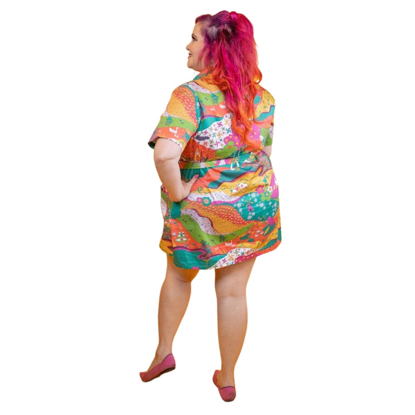 A model wearing a stretch cotton mini dress with a rounded wide collar, short sleeves, and a bright purple plastic zipper running down the length of the dress. It has an intricate multicolored psychedelic pattern including pinwheels, apples, hands, birds, and other illustrations in pink, yellow, orange, blue, green, and white. Dress comes with a matching sash style belt. Shown from back