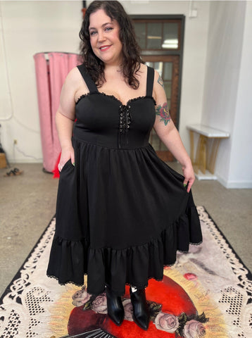 A model wearing a sleeveless black ponte knit swing dress with silver lace up eyelet detail at the sweetheart bodice. Black floral lace trim at the bodice and ruffled skirt. Shown from the front