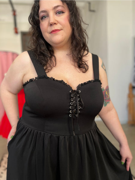 A model wearing a sleeveless black ponte knit swing dress with silver lace up eyelet detail at the sweetheart bodice. Black floral lace trim at the bodice and ruffled skirt. Shown from the front in close up