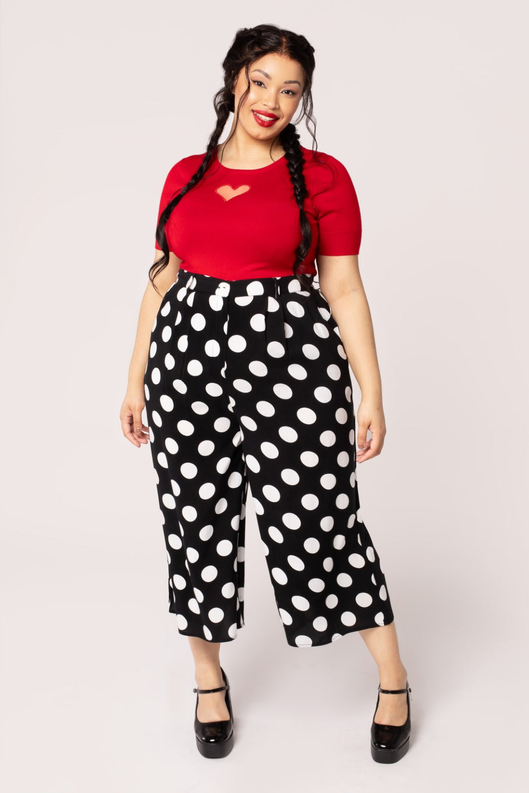 A plus size model wearing a pair of cropped and high waisted rayon trousers in a large black and white polka dot pattern. They have pleats and a zip & button front. Shown from front