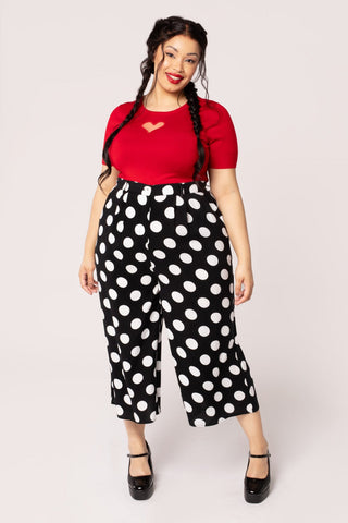 A plus size model wearing a pair of cropped and high waisted rayon trousers in a large black and white polka dot pattern. They have pleats and a zip & button front. Shown from front