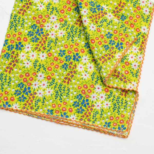 Cotton seersucker lime green square scarf with all over wildflower pattern in white, red, green, and blue. Has an orange embroidered scalloped trim. Shown in close up