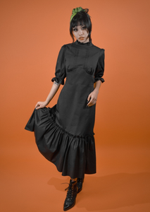 A model wearing a black midi length dress with a high ruffled collar, slightly puffed shoulders, elbow length sleeves with elasticized cuffs, pintuck detail down the center of the bodice, and a shaped gathered underbust seam. The skirt has a gathered ruffle and extra ruffle at the hem. Shown from the front