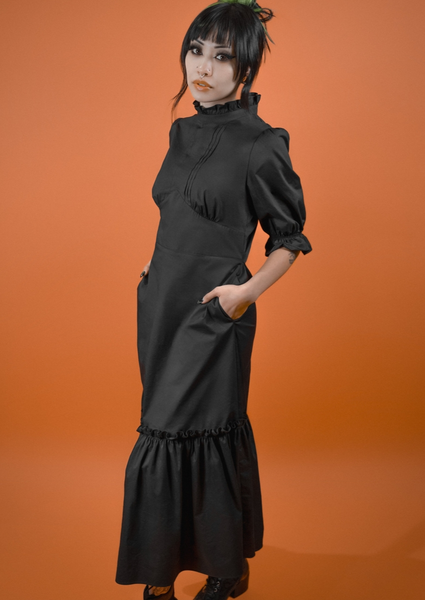 A model wearing a black midi length dress with a high ruffled collar, slightly puffed shoulders, elbow length sleeves with elasticized cuffs, pintuck detail down the center of the bodice, and a shaped gathered underbust seam. The skirt has a gathered ruffle and extra ruffle at the hem. Shown from the front at a three quarter angle with hands in pockets