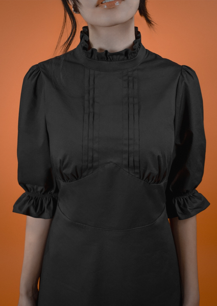 A model wearing a black midi length dress with a high ruffled collar, slightly puffed shoulders, elbow length sleeves with elasticized cuffs, pintuck detail down the center of the bodice, and a shaped gathered underbust seam. The skirt has a gathered ruffle and extra ruffle at the hem. Shown from the front in close up of the bodice