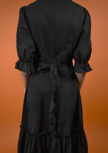 A model wearing a black midi length dress with a high ruffled collar, slightly puffed shoulders, elbow length sleeves with elasticized cuffs, pintuck detail down the center of the bodice, and a shaped gathered underbust seam. The skirt has a gathered ruffle and extra ruffle at the hem. Shown from the back with waist tie in a bow 