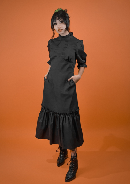 A model wearing a black midi length dress with a high ruffled collar, slightly puffed shoulders, elbow length sleeves with elasticized cuffs, pintuck detail down the center of the bodice, and a shaped gathered underbust seam. The skirt has a gathered ruffle and extra ruffle at the hem. Shown from the front