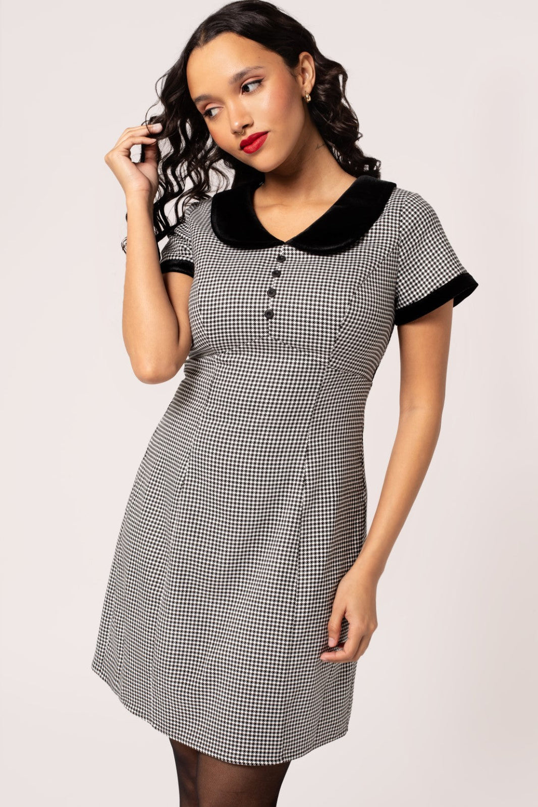 A model wearing a short sleeve mini  dress in a black and white micro houndstooth print. It has a rounded black velvet collar and black velvet sleeve cuffs. There are round black plastic buttons down the bodice and the skirt is slightly a-line and above the knee. Shown from the front in close up