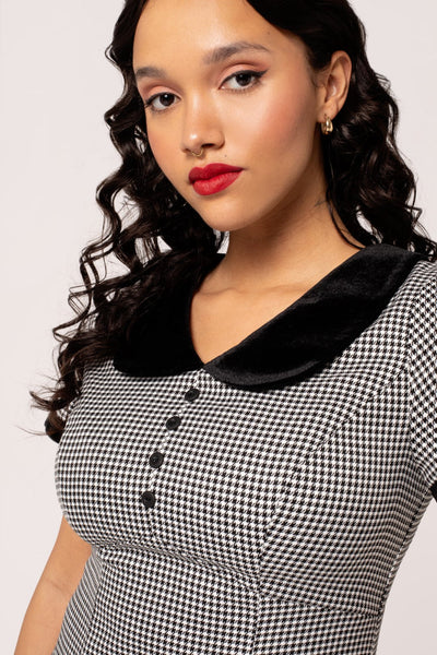 A model wearing a short sleeve mini dress in a black and white micro houndstooth print. It has a rounded black velvet collar and black velvet sleeve cuffs. There are round black plastic buttons down the bodice and the skirt is slightly a-line and above the knee. Shown from the bodice up in close up 