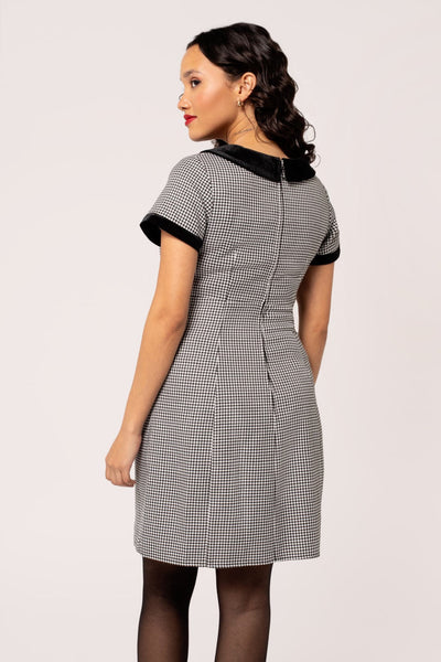 A model wearing a short sleeve mini dress in a black and white micro houndstooth print. It has a rounded black velvet collar and black velvet sleeve cuffs. There are round black plastic buttons down the bodice and the skirt is slightly a-line and above the knee. Shown from the back