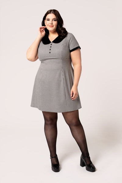 A plus size model wearing a short sleeve mini dress in a black and white micro houndstooth print. It has a rounded black velvet collar and black velvet sleeve cuffs. There are round black plastic buttons down the bodice and the skirt is slightly a-line and above the knee. Shown from the front