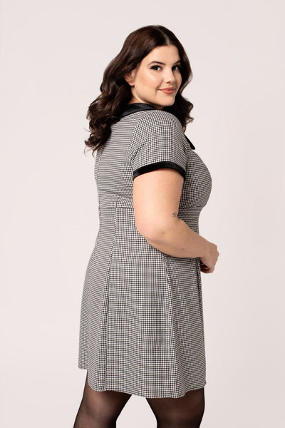 A plus size model wearing a short sleeve mini dress in a black and white micro houndstooth print. It has a rounded black velvet collar and black velvet sleeve cuffs. There are round black plastic buttons down the bodice and the skirt is slightly a-line and above the knee. Shown from the back