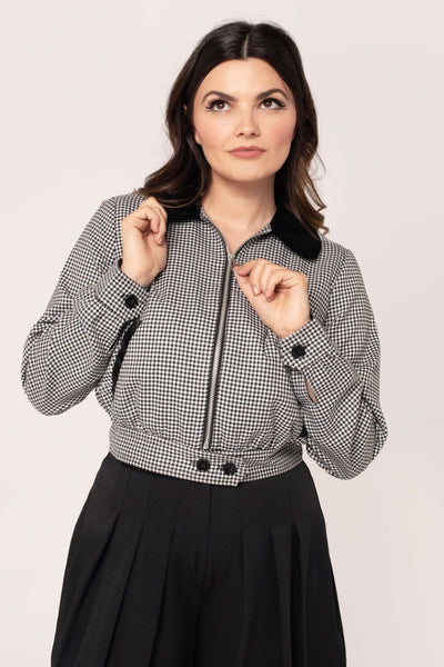 model wearing a black and white micro houndstooth print bomber-style jacket with rounded black velvet collar and front slash pockets with black velvet trim, o-ring front zipper, and black plastic buttons at bottom of waistband. Shown in close up with jacket zipped up from front