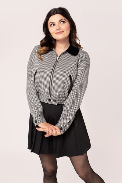 model wearing a black and white micro houndstooth print bomber-style jacket with rounded black velvet collar and front slash pockets with black velvet trim, o-ring front zipper, and black plastic buttons at bottom of waistband. Shown with jacket zipped up from front