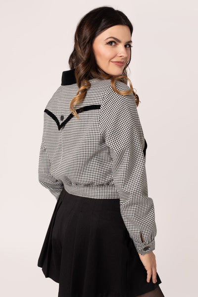 model wearing a black and white micro houndstooth print bomber-style jacket with rounded black velvet collar and front slash pockets with black velvet trim, o-ring front zipper, and black plastic buttons at bottom of waistband, and back yoke detail with velvet trim. Shown from back.