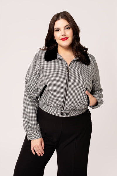 a plus size model wearing a black and white micro houndstooth print bomber-style jacket with rounded black velvet collar and front slash pockets with black velvet trim, o-ring front zipper, and black plastic buttons at bottom of waistband. Shown with jacket zipped up from front