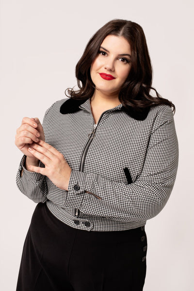 a plus size model wearing a black and white micro houndstooth print bomber-style jacket with rounded black velvet collar and front slash pockets with black velvet trim, o-ring front zipper, and black plastic buttons at bottom of waistband. Shown in close up with jacket zipped up from front