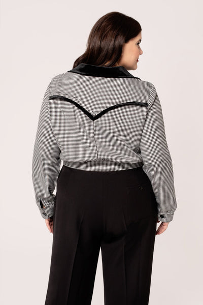 a plus size model wearing a black and white micro houndstooth print bomber-style jacket with rounded black velvet collar and front slash pockets with black velvet trim, o-ring front zipper, and black plastic buttons at bottom of waistband, and back yoke detail with velvet trim. Shown from back.