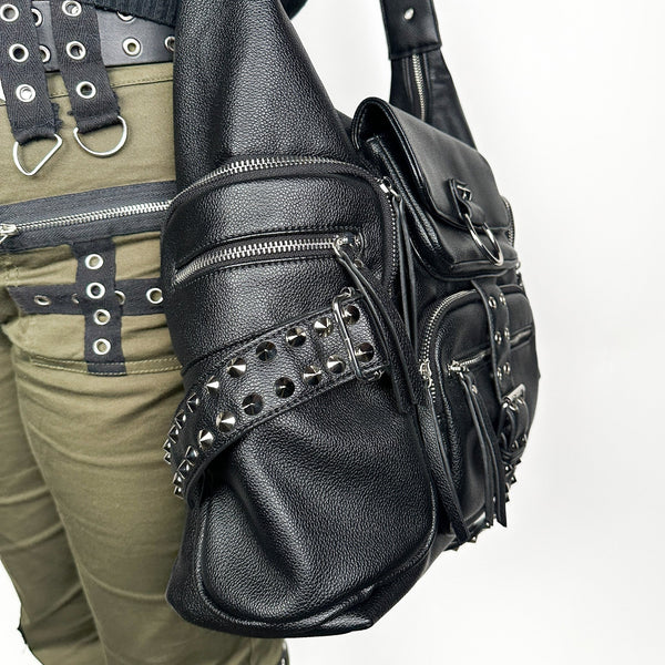 black textured faux leather crossbody bag with 8 exterior pockets with silver metal zip closure and cone stud & o-ring detail on oversized strap detail. Bag has 27" adjustable self strap. Shown from side in close up worn by model