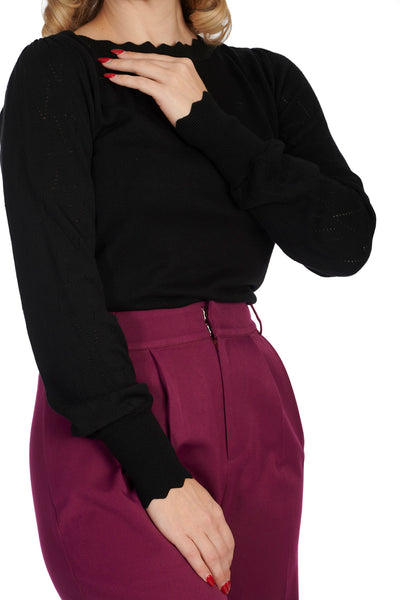 Model wearing long sleeved black knit sweater with scalloped slash neckline, slightly puffed shoulders, geometric pointelle details at the sleeves, and scalloped cuffs and bottom band. Shown from the side