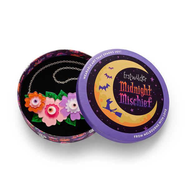 A colorful floral necklace with four stylized flowers in orange, pink, and purple, all with eyeball centers, and accented with green leaves and a silver chain, shown in illustrated round gift box packaging