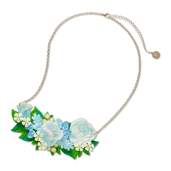 A floral statement necklace featuring light blue and white roses, green leaves, and small white flowers on a delicate gold chain, shown against a plain white background.