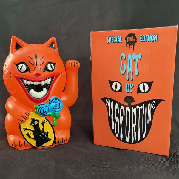 An orange maneki-neko cat figurine meant to resemble the cat Blanche from the movie House, with a wide grin, white eyes, and one raised paw, holding blue roses and a yellow sign with a black cat silhouette, photographed next to its gift box against a dark gray background.