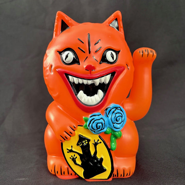 An orange maneki-neko cat figurine meant to resemble the cat Blanche from the movie House, with a wide grin, white eyes, and one raised paw, holding blue roses and a yellow sign with a black cat silhouette, photographed against a dark gray background.