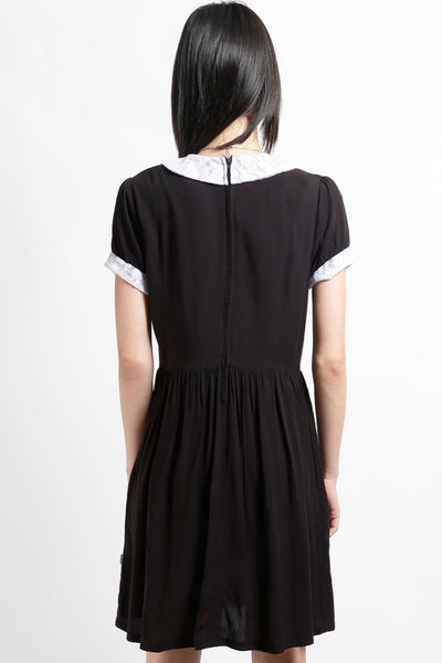 model wearing black rayon shirtwaist dress with white lace peter pan collar, matching lace cuffs at short sleeves, round black plastic buttons at bodice and gathered above the knee skirt. Shown from back