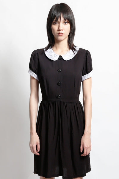 model wearing black rayon shirtwaist dress with white lace peter pan collar, matching lace cuffs at short sleeves, round black plastic buttons at bodice and gathered above the knee skirt. Shown from front in close up