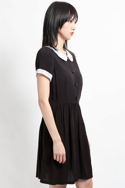 model wearing black rayon shirtwaist dress with white lace peter pan collar, matching lace cuffs at short sleeves, round black plastic buttons at bodice and gathered above the knee skirt. Shown from side