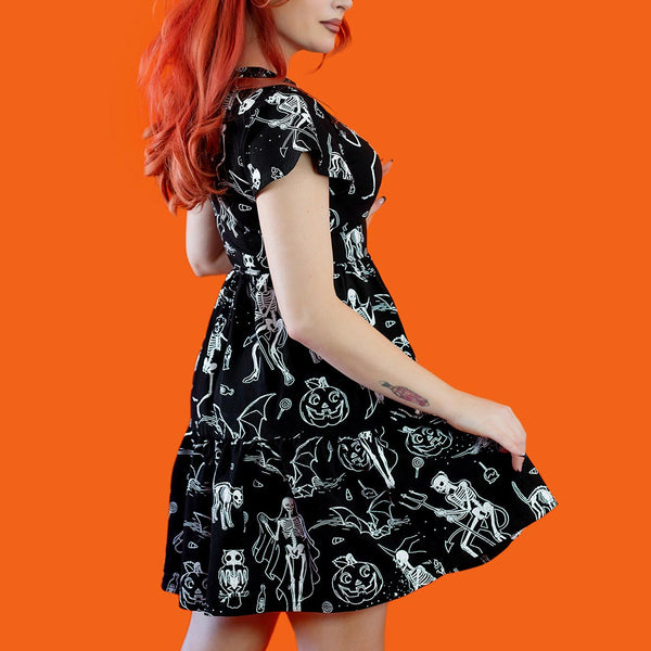 Model wearing a black cotton jersey knit dress with white skeleton, owl, pumpkin, witch and bat print. Dress has a shallow v-neckline, short flutter sleeves, a knee length double tiered skirt with ruffle hem and pockets. Shown from back against an orange background
