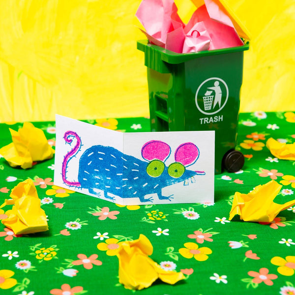 heavyweight card stock note card with a neon pink, green, and blue risograph image of a cartoony rat that wraps around both exterior sides of card. Shown on green and yellow floral background in front of mini trash can surrounded by crumpled paper