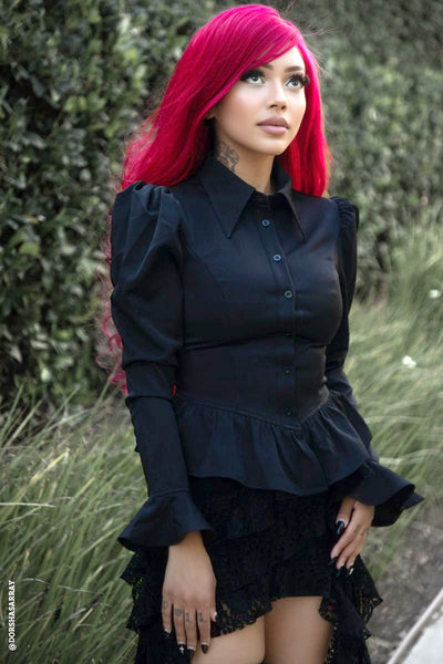 model wearing black chiffon long sleeve blouse with button-down front, pointed collar, exaggerated puffed shoulders, ruffled cuffs, and peplum style ruffled gathered bottom hem. Shown from side