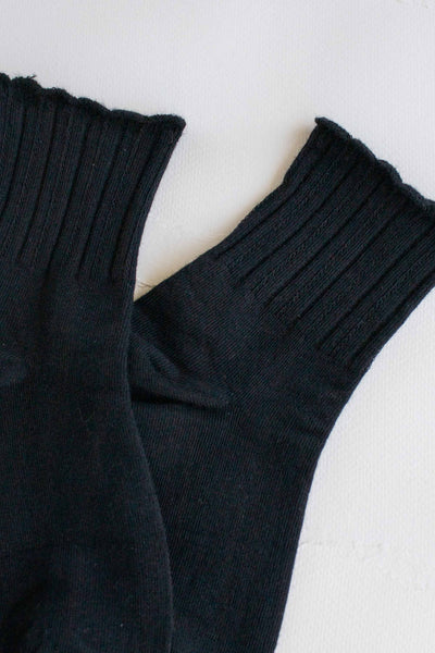 Black cotton knit socks with ribbed pattern and scalloped cuffs. Shown flat in close up