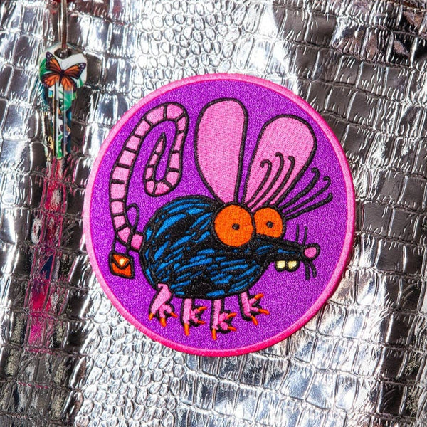 round embroidered patch with purple background and pink border of black and blue cartoon rat with big pink ears, red eyes, a curly tail with red purse hanging from it, and long red pointy nails. Shown on silver metallic snakeskin background