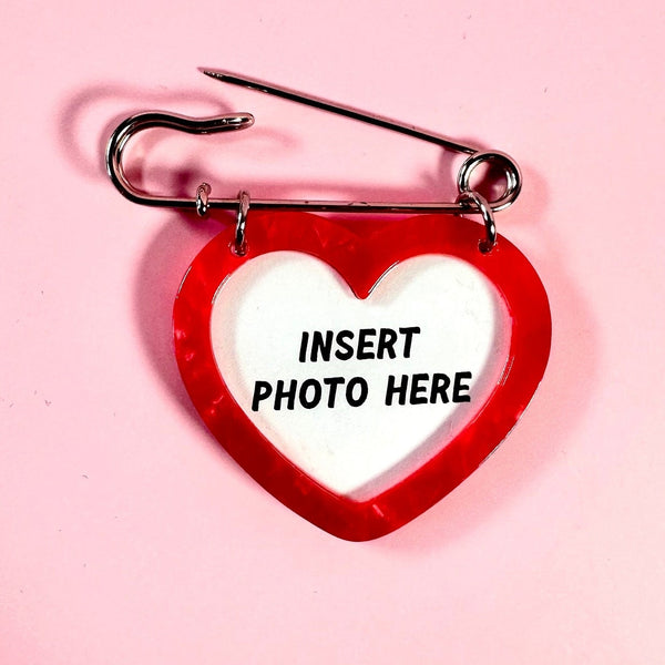 A safety pin brooch with an attached red marbled heart pendant and a white center that reads 'INSERT PHOTO HERE' in black text, photographed on a solid pink background.
