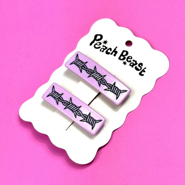 set of two baby pink rectangular acetate barrettes with French style closure and carved black detail of three links of barbed wire. Shown on backing card on pink background