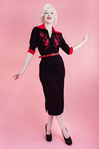 model wearing black sweater knit dress with bright red rolled collar, v-neckline, elbow length sleeves with red cuffs, and embroidered detail across the bodice in matching red of heart-shaped lock, key, and gloved hand. Dress has pencil skirt with side slit and red patent faux leather skinny belt. Shown from front