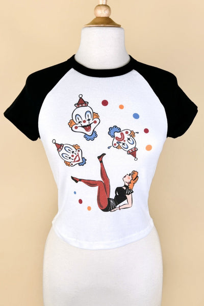 white crop t-shirt with black raglan sleeves & collar and printed illustration of woman in circus acrobat costume lying on her back with legs posed in the air surrounded by three large primary colored clown heads and primary colored dots. Shown on dress form