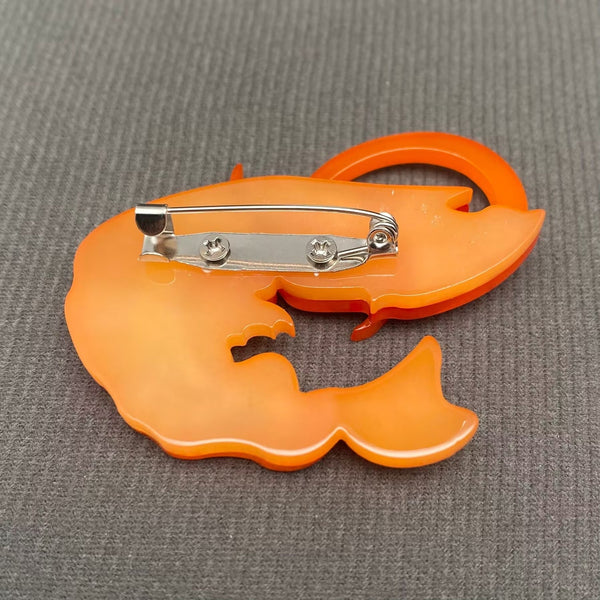 layered acrylic resin brooch of an orange marbled shrimp seen from the side with single black rhinestone jewel for an eye. Shown flat from back